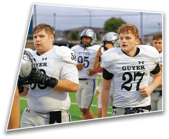 A group of adults wearing white football uniforms with the word Guyer and numbers on them are standing and walking on a sports field. Description generated by AI
