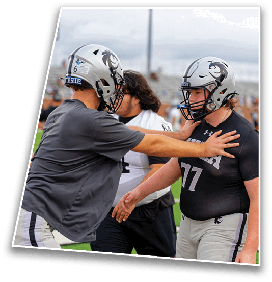Two people wearing football helmets and athletic gear practice blocking techniques on a sports field while a third person observes in the background. Description generated by AI