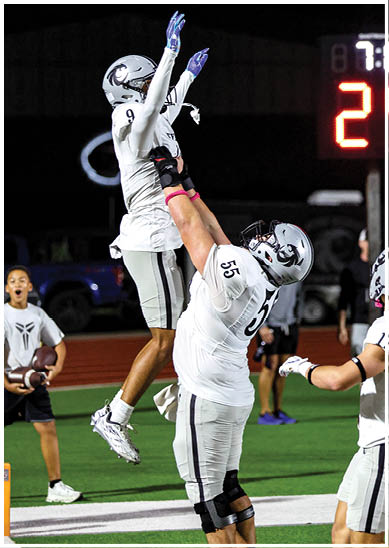 Two people in football uniforms are celebrating on a field with one person lifting the other while a scoreboard is visible in the background. Description generated by AI