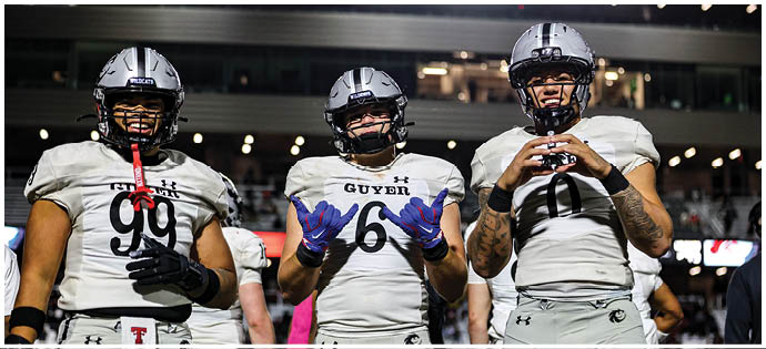 Three people wearing light gray sports uniforms stand side by side on a field with a crowd in the background. Description generated by AI