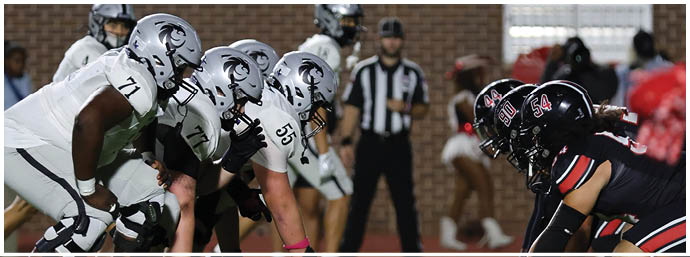 Two opposing American football teams are lined up at the line of scrimmage preparing for a play on a field with a blurred referee and cheerleaders in the background. Description generated by AI