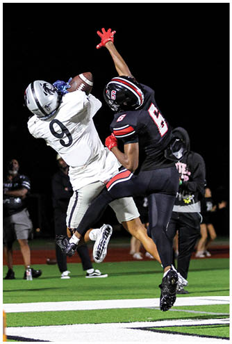 Two people in football uniforms are jumping and reaching for a football near the end zone on a field at night. Description generated by AI