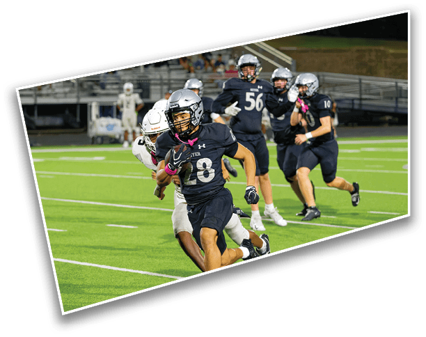 A person in a black football uniform is running with the ball while being chased by a person in a white uniform on a green field with other players and empty bleachers in the background. Description generated by AI