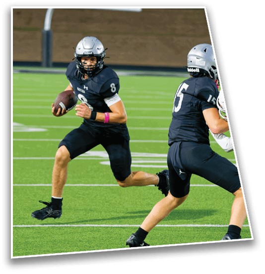 Two people in black football uniforms are playing on a green field, with one holding a football and running while the other appears to be moving to block or assist. Description generated by AI
