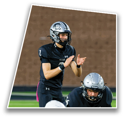 A person in a black football uniform and helmet is clapping hands while another person in a similar uniform and helmet is crouched in front on a football field. Description generated by AI
