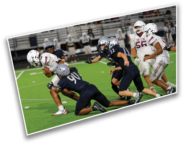 People playing a football game on a green field with some players in white uniforms and others in dark uniforms actively engaged in a tackle. Description generated by AI