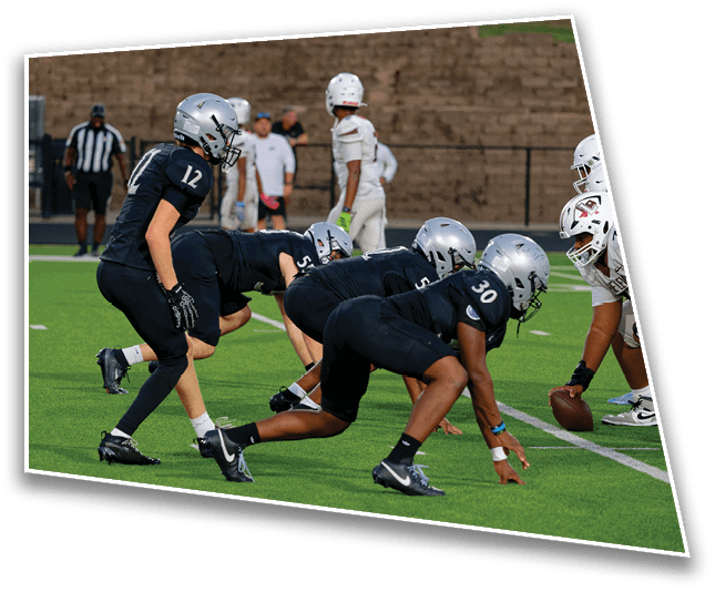 Several football players in black uniforms and silver helmets are lined up on a green field facing opponents in white uniforms, preparing for a play. Description generated by AI