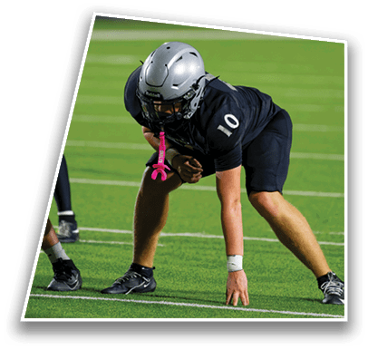 A person wearing a silver helmet and black sports uniform with the number 10 is crouched on a green football field, preparing for a play. Description generated by AI