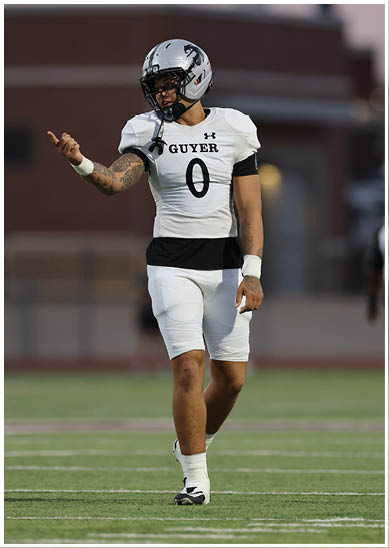 A person wearing a white football uniform with the number zero and a helmet is standing on a football field making a hand gesture. Description generated by AI