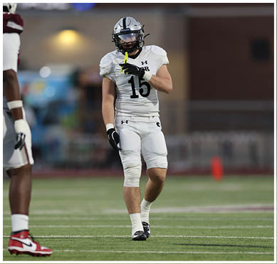 A person in a white football uniform with the number 15 is standing on a field holding a yellow marker near their mouth while another person in a maroon and white uniform is partially visible. Description generated by AI