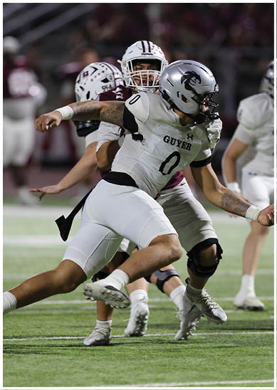 A person wearing a white football uniform with the word “GUYER" and the number zero is running on a field while being closely pursued by another person in a maroon and white uniform. Description generated by AI