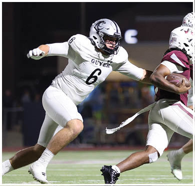 A person in a white football uniform is reaching out to tackle a person in a maroon football uniform who is holding the ball while another person in a maroon uniform runs ahead on a lit football field at night. Description generated by AI
