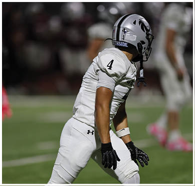 A person wearing a white football uniform and helmet with the number 4 is on a field with blurred players in the background. Description generated by AI