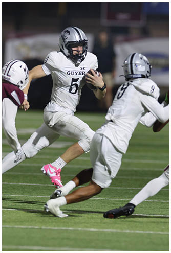 Four people in football uniforms are actively engaged in a play on a green field under stadium lights, with two wearing white uniforms and two wearing maroon uniforms. Description generated by AI
