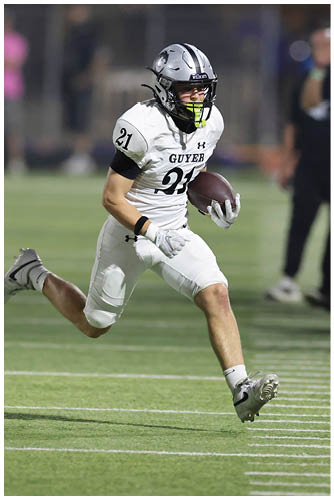 A person wearing a white football uniform is running on a field while holding a football near the sideline with blurred people standing in the background. Description generated by AI