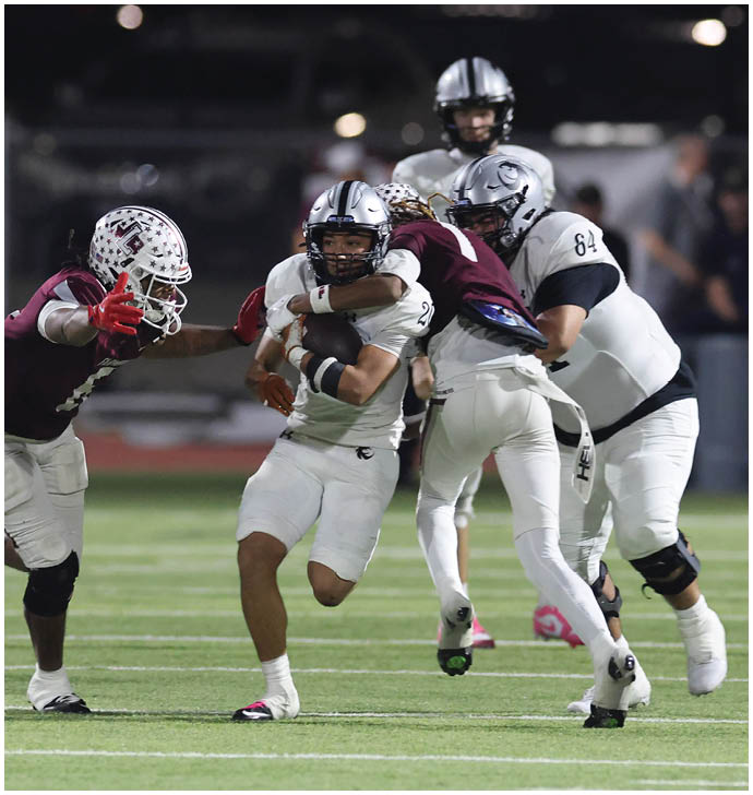 Four people in football uniforms are engaged in a play on a field, with one person in white holding the ball while two others in maroon attempt to tackle or block. Description generated by AI