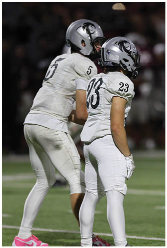 Two people wearing white football uniforms and helmets stand on a field, one facing sideways with the number 23 on the jersey and the other partially visible with the number 5. Description generated by AI