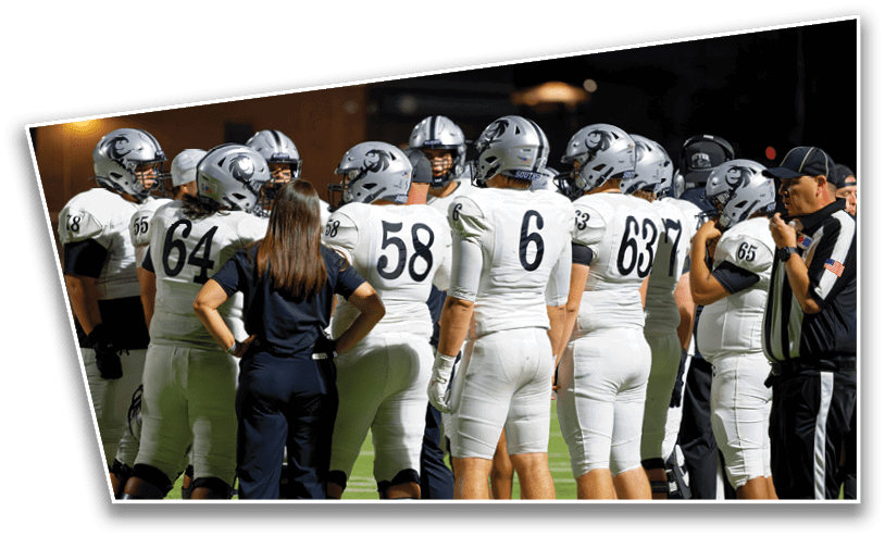 A group of people in white football uniforms and helmets are gathered on a field with two individuals in dark clothing and one person in a referee uniform standing nearby. Description generated by AI