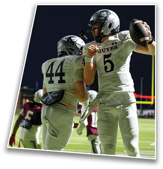 Two people in white football uniforms celebrating on a field at night while other players in dark uniforms stand in the background. Description generated by AI