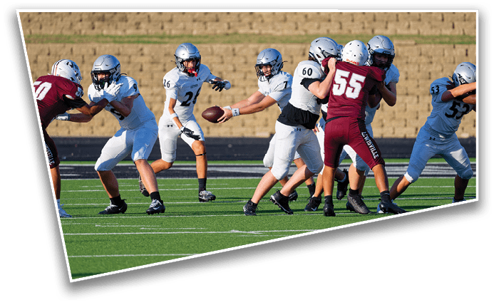 Players in white and maroon football uniforms are engaged in a play on a green field with a brick wall in the background. Description generated by AI