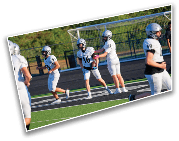 Several people in football uniforms and helmets are on a field near a goalpost, with one person holding a football and another reaching out to them. Description generated by AI