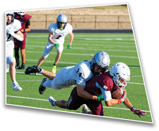 A group of people in football uniforms are playing on a field with one person in white tackling another person in maroon who is holding a football. Description generated by AI