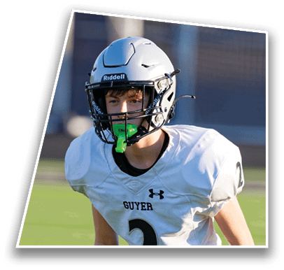 A person wearing a white football uniform with the word “GUYER" and a helmet with a green mouthguard is on a grassy field. Description generated by AI