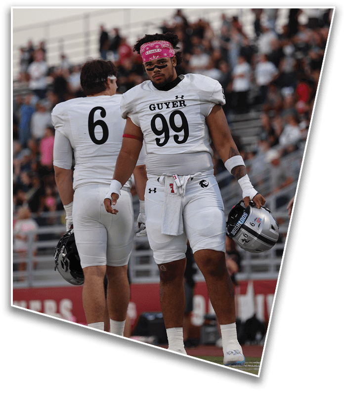 Two people in white football uniforms with numbers 99 and 6 stand on a field with a crowd in the background, with the person wearing number 99 holding a helmet and wearing a pink headband. Description generated by AI