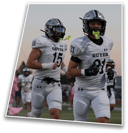 Two people in white football uniforms with helmets and mouthguards are running on a field with others and a pink flag in the background. Description generated by AI