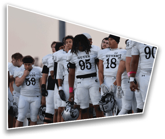 A group of people wearing white football jerseys with the word “GUYER" stand closely together outdoors during dusk. Description generated by AI