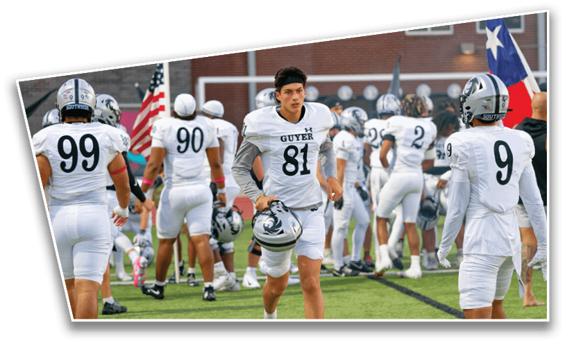 A group of people in white football uniforms with helmets are gathered on a field, with one person holding a helmet and walking toward the camera. Description generated by AI
