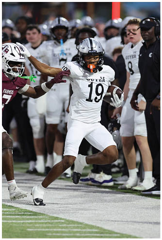A person wearing a white football uniform with the number 19 is running with the ball while another person in a maroon uniform attempts to grab their helmet on the sideline during a game. Description generated by AI