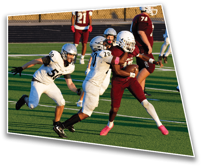 People in football uniforms are actively playing on a green field with one person in a maroon uniform holding the ball while being tackled by two people in white uniforms. Description generated by AI