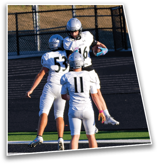 Three people wearing football helmets and white jerseys are engaged in a play on a field with a chain-link fence in the background. Description generated by AI