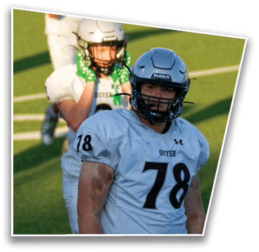 Two people wearing football helmets and white jerseys with the word “GUYER" are on a grassy field, with one person in the foreground and the other adjusting their helmet in the background. Description generated by AI
