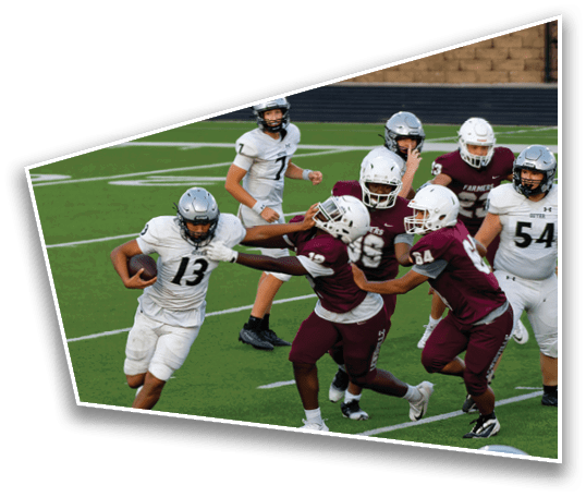 A person in a white football uniform is running with the ball while three people in maroon uniforms attempt to block or tackle them on a green football field. Description generated by AI