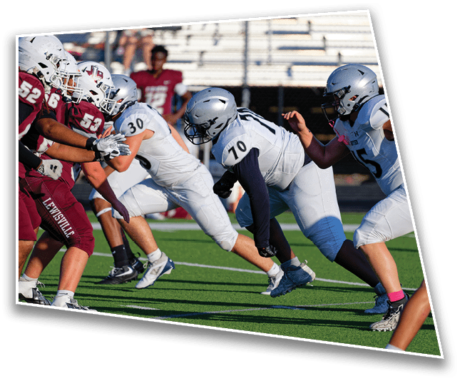 Several people in football uniforms and helmets are engaged in a play on a green field with one person in a white uniform lunging forward. Description generated by AI