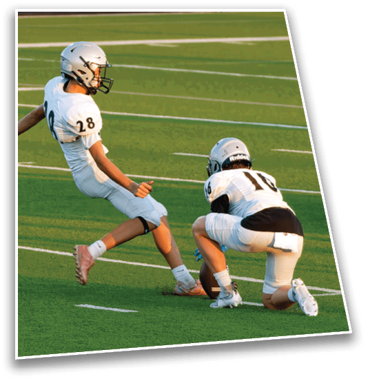 Two people in football uniforms are on a field with one holding the ball on the ground while the other prepares to kick it. Description generated by AI