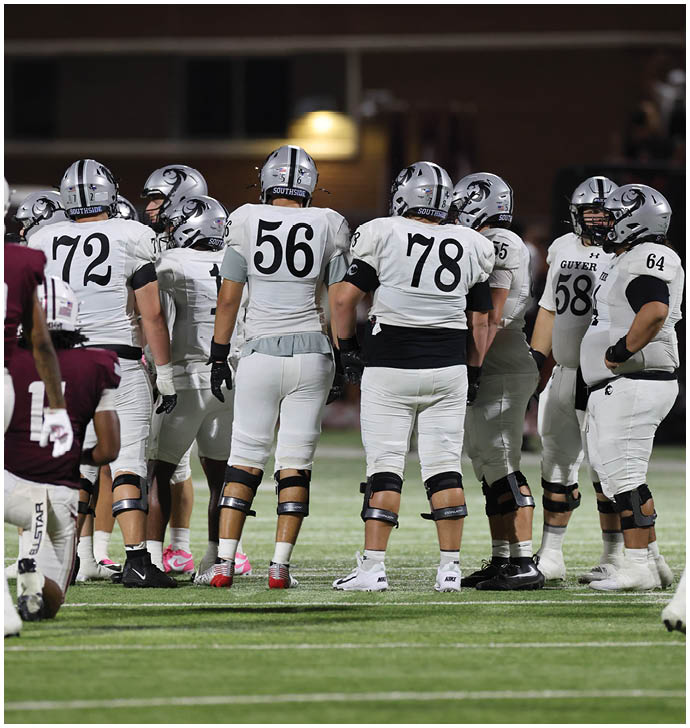 A group of football players in white uniforms and helmets are standing together on a field during a game or practice. Description generated by AI