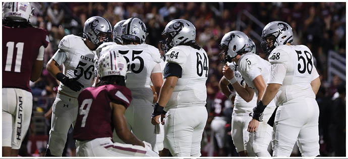A group of football players in white uniforms and helmets are huddled together on a field with a crowd in the background. Description generated by AI