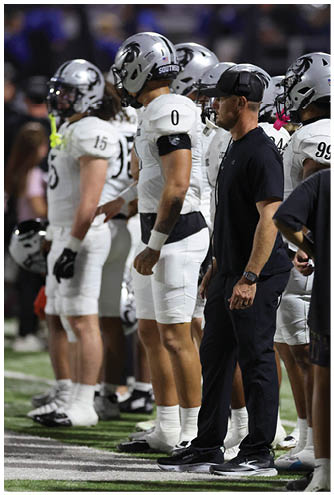 Several people in football uniforms and helmets stand on the sidelines of a field next to a person in a black outfit and cap. Description generated by AI
