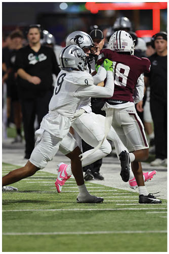 Two football players in white and maroon uniforms compete for the ball near the sideline during a nighttime game with spectators and team members in the background. Description generated by AI