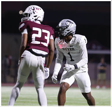 Two people wearing football uniforms and helmets are on a field at night, with one person in a white uniform facing the other in a maroon uniform. Description generated by AI