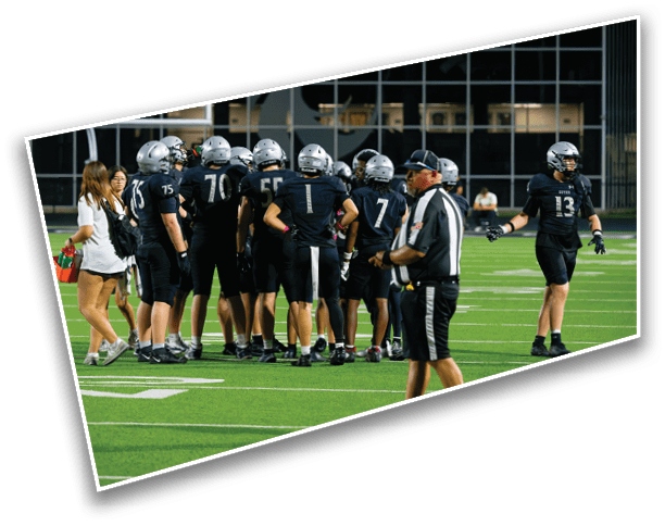 A group of people wearing football uniforms and helmets are gathered on a field with two adults standing nearby, one holding water bottles. Description generated by AI
