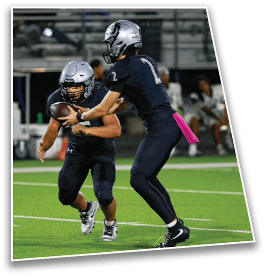 Two people wearing football helmets and uniforms are on a field with one handing a football to the other during a game or practice. Description generated by AI