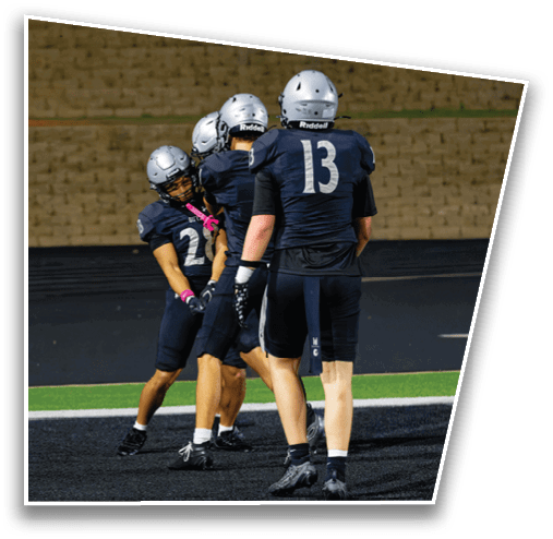 Three people wearing football helmets and dark uniforms are standing on a field near an end zone with a scoreboard showing time and score in the background. Description generated by AI