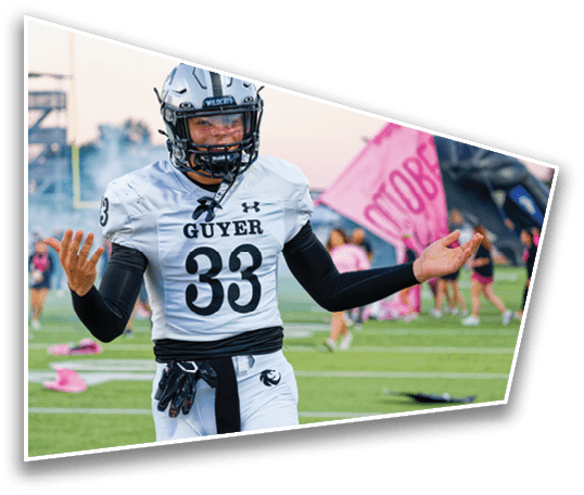 A person in a football uniform with the number 33 and the word “GUYER" on the jersey stands on a field with arms raised in a questioning or shrugging gesture while cheerleaders and a pink banner are visible in the background. Description generated by AI