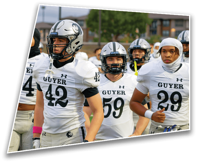 A group of people wearing white football uniforms with helmets and numbers stand together on a field with a blurred background of trees and buildings. Description generated by AI