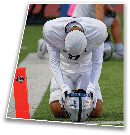 A person in white football gear kneels on the field holding a helmet with the word “SOUTHSIDE" on it near an orange marker with a Texas logo. Description generated by AI