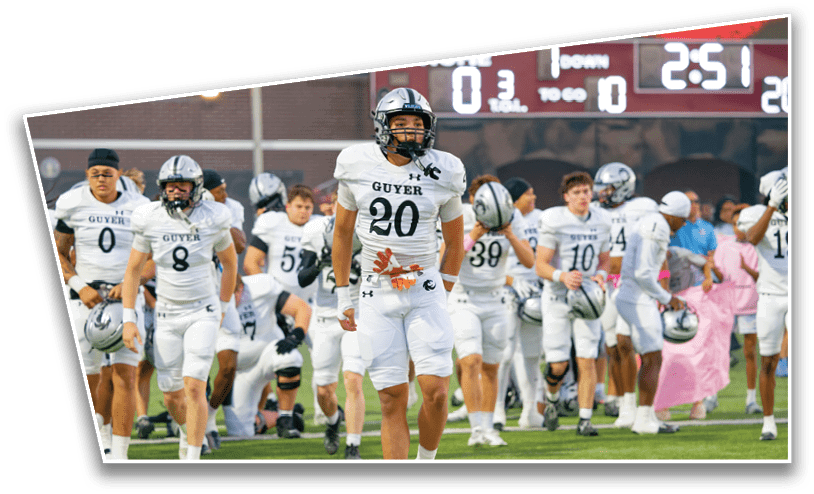 A group of people wearing white football uniforms with “Guyer" on the jerseys are walking on a green field, some holding helmets. Description generated by AI